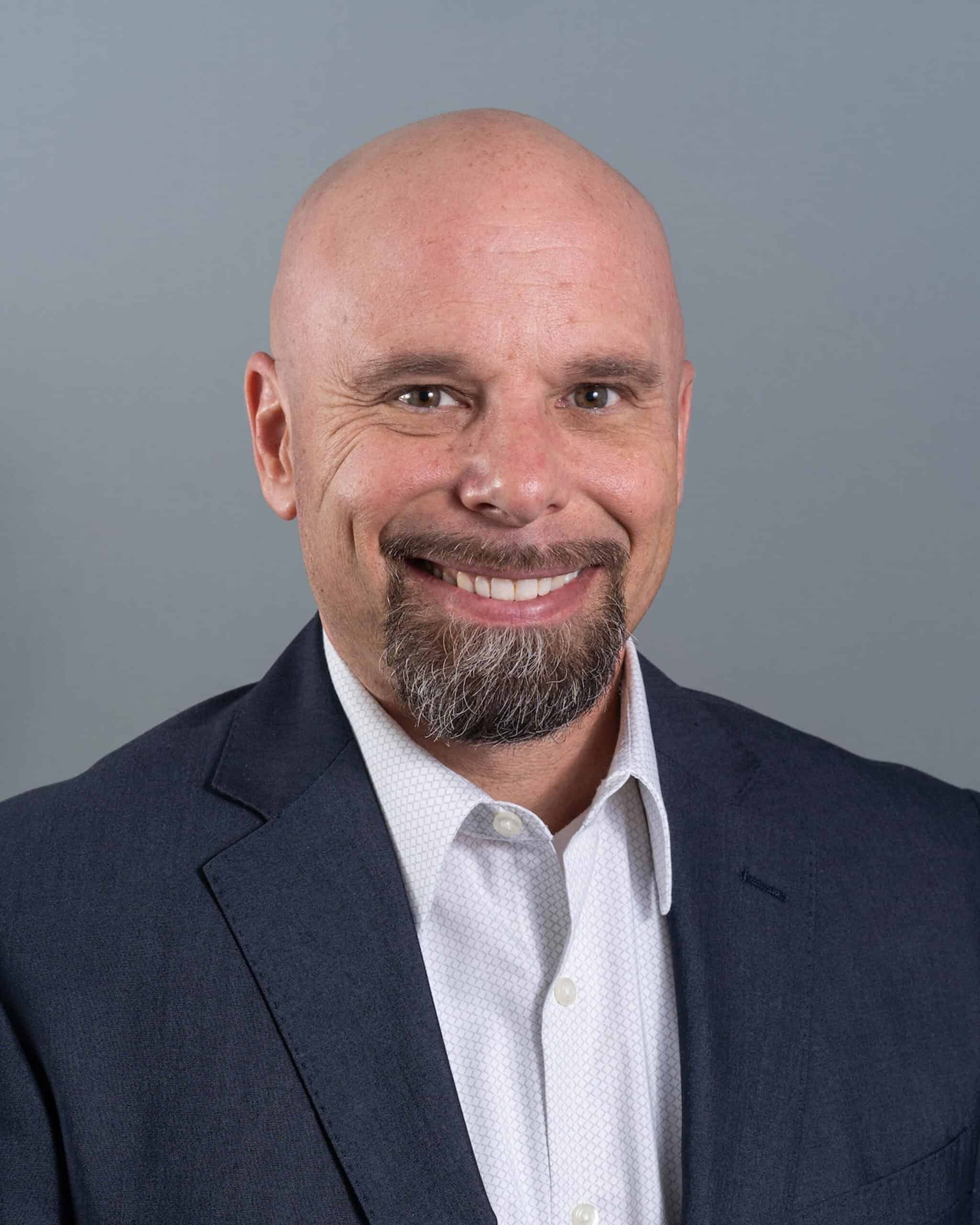Smiling bald man in a suit and white shirt against gray background.