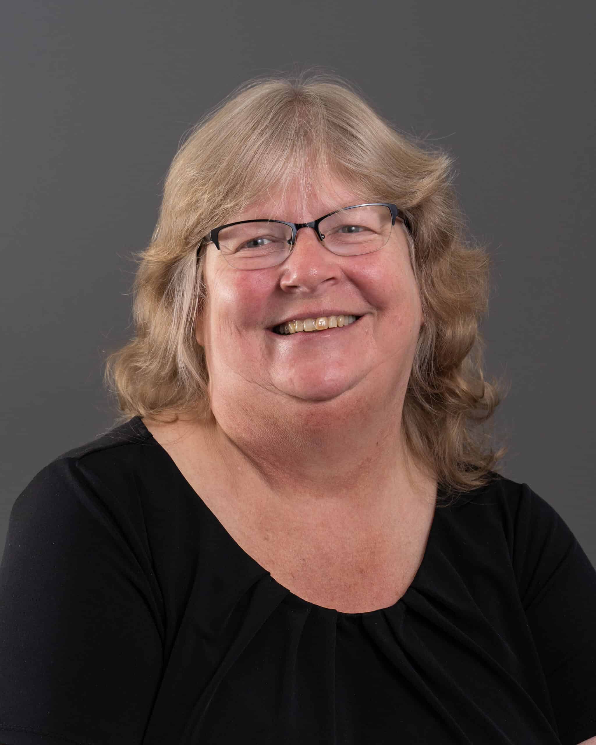 Smiling woman with glasses and blonde hair in a black top against gray background.