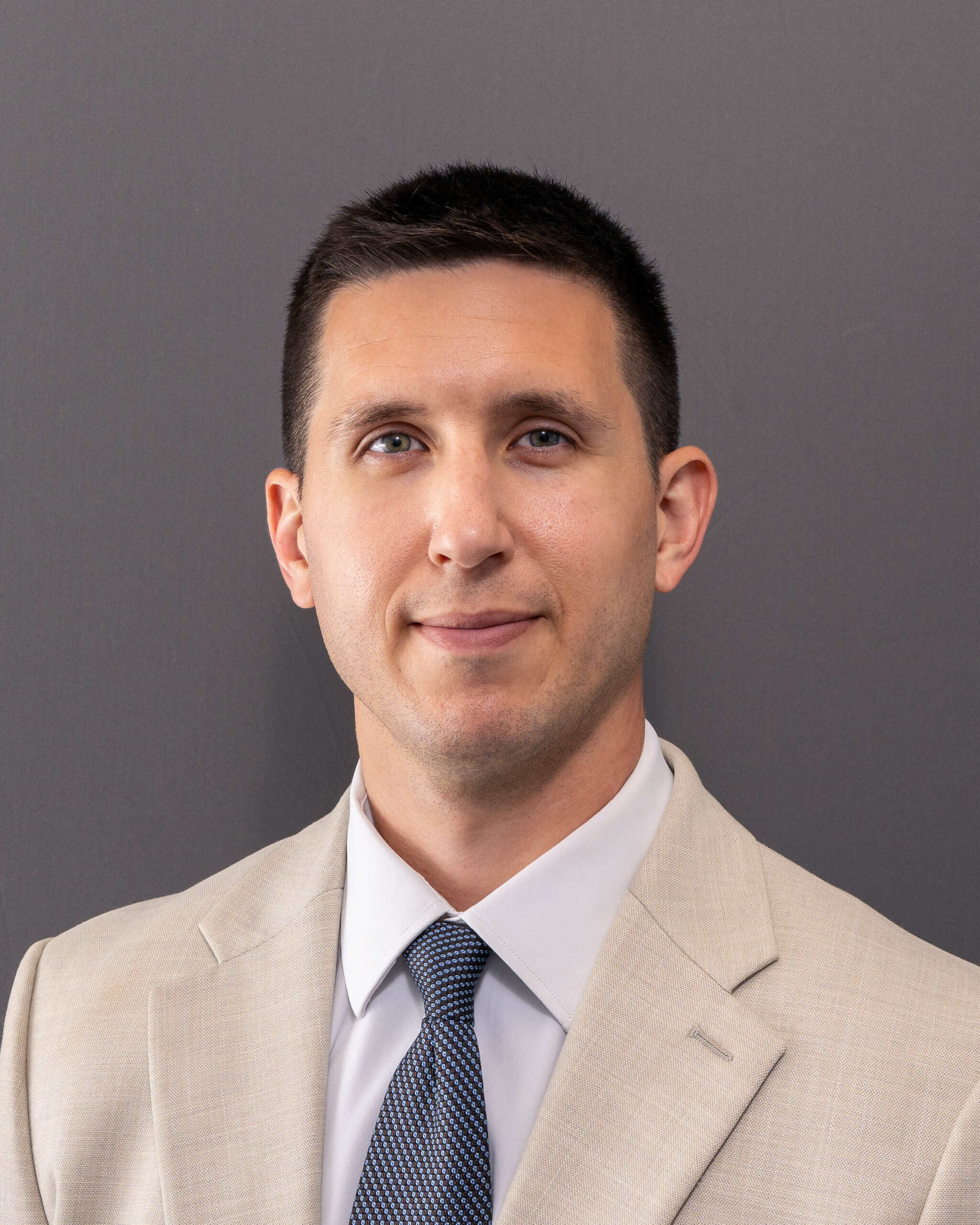 Professional man in beige suit and blue tie against gray background.