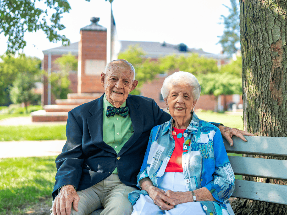 Elderly man and woman smiling on park bench with trees and building in background.