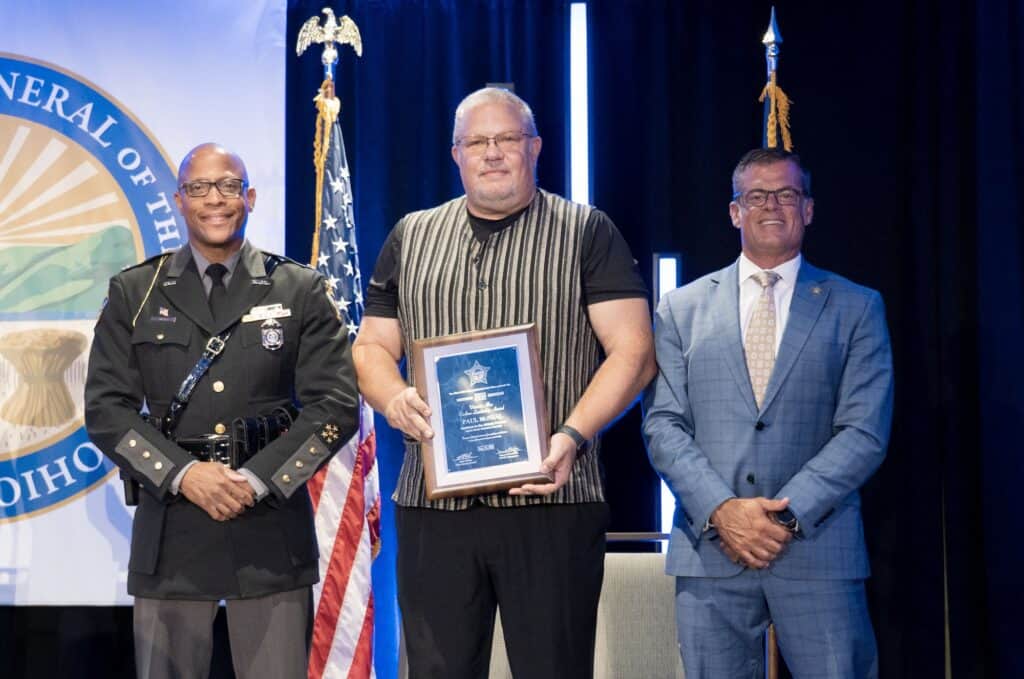 Three men on stage, one holding a framed award, with flags and a banner in the background.