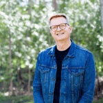 Man smiling in denim jacket outdoors with trees in background.