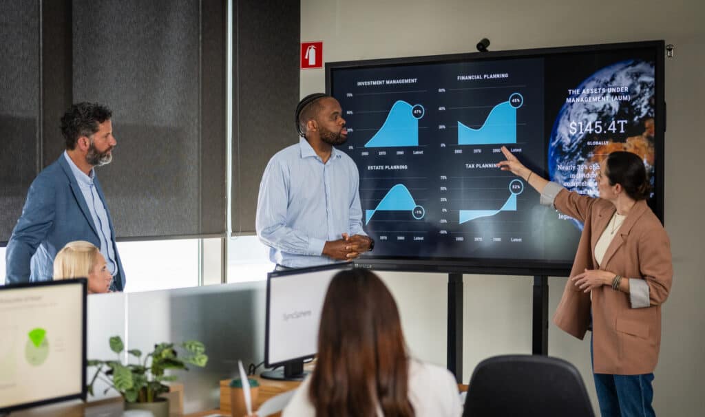 A team leader walks colleagues through performance data on a large display in a busy office, showing how MVNU's online Master of Organizational Leadership builds the skills to guide teams, communicate clearly, and lead change with integrity.