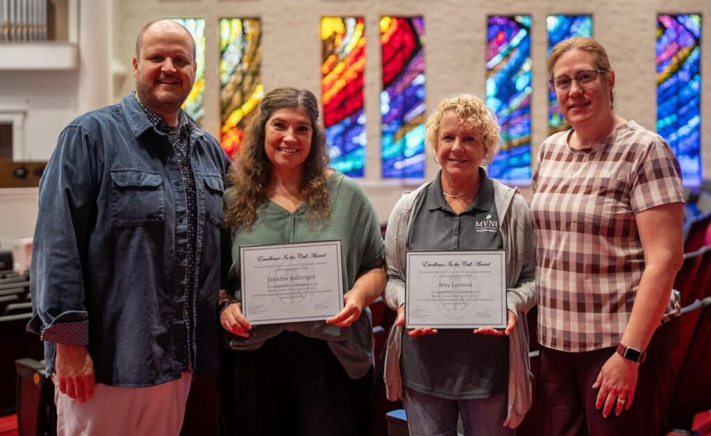 Four people standing inside a church with stained glass windows, two holding certificates.