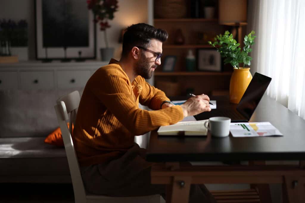 A man takes notes beside his laptop at a dining table, researching which MBA concentration fits his career goals.