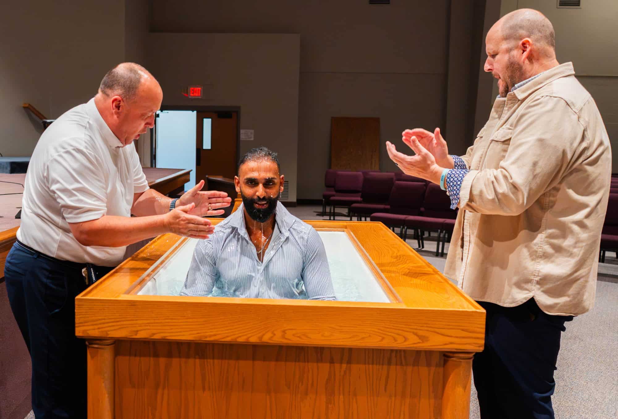 Three men in a baptismal pool, one seated and two standing, inside a church setting.