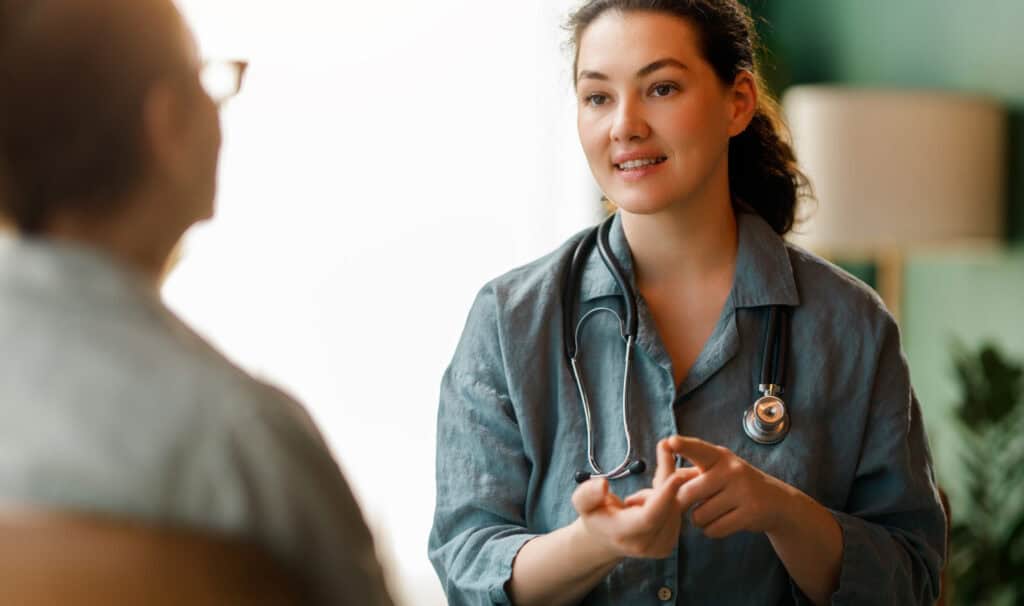 A nurse practitioner with a stethoscope talking with a patient during a seated consultation, reflecting the care choices behind the PMHNP vs FNP decision.