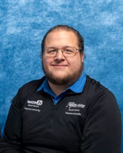 Portrait of Professor Micah Andrew Gerber with glasses and beard, smiling, against blue background.
