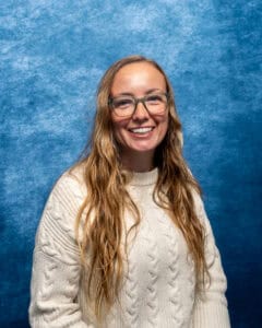 Smiling woman with long wavy hair and glasses against blue background.