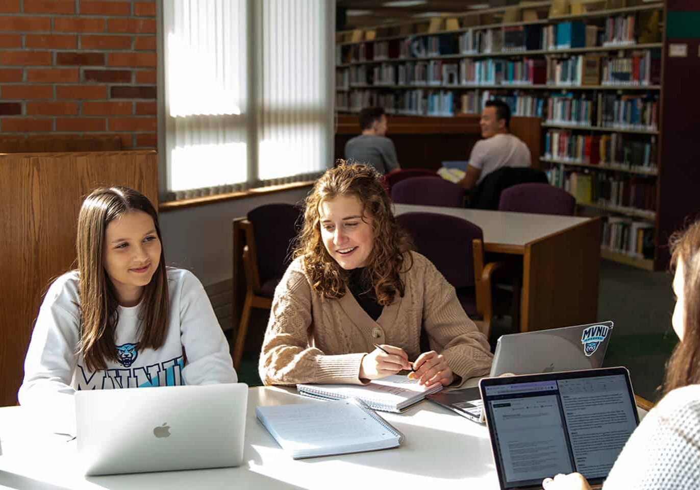 Students studying and collaborating at a library table with laptops and books.