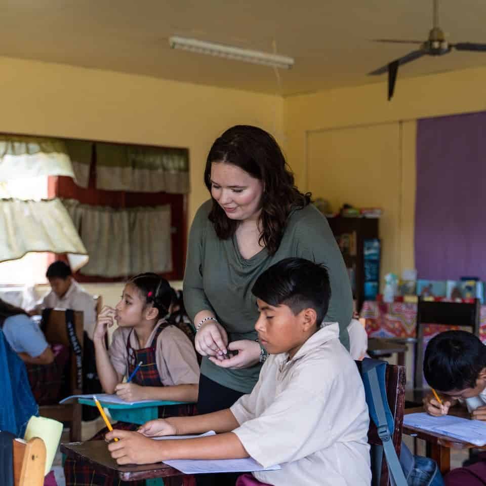 A teacher assisting students in a classroom with laptops and notebooks.