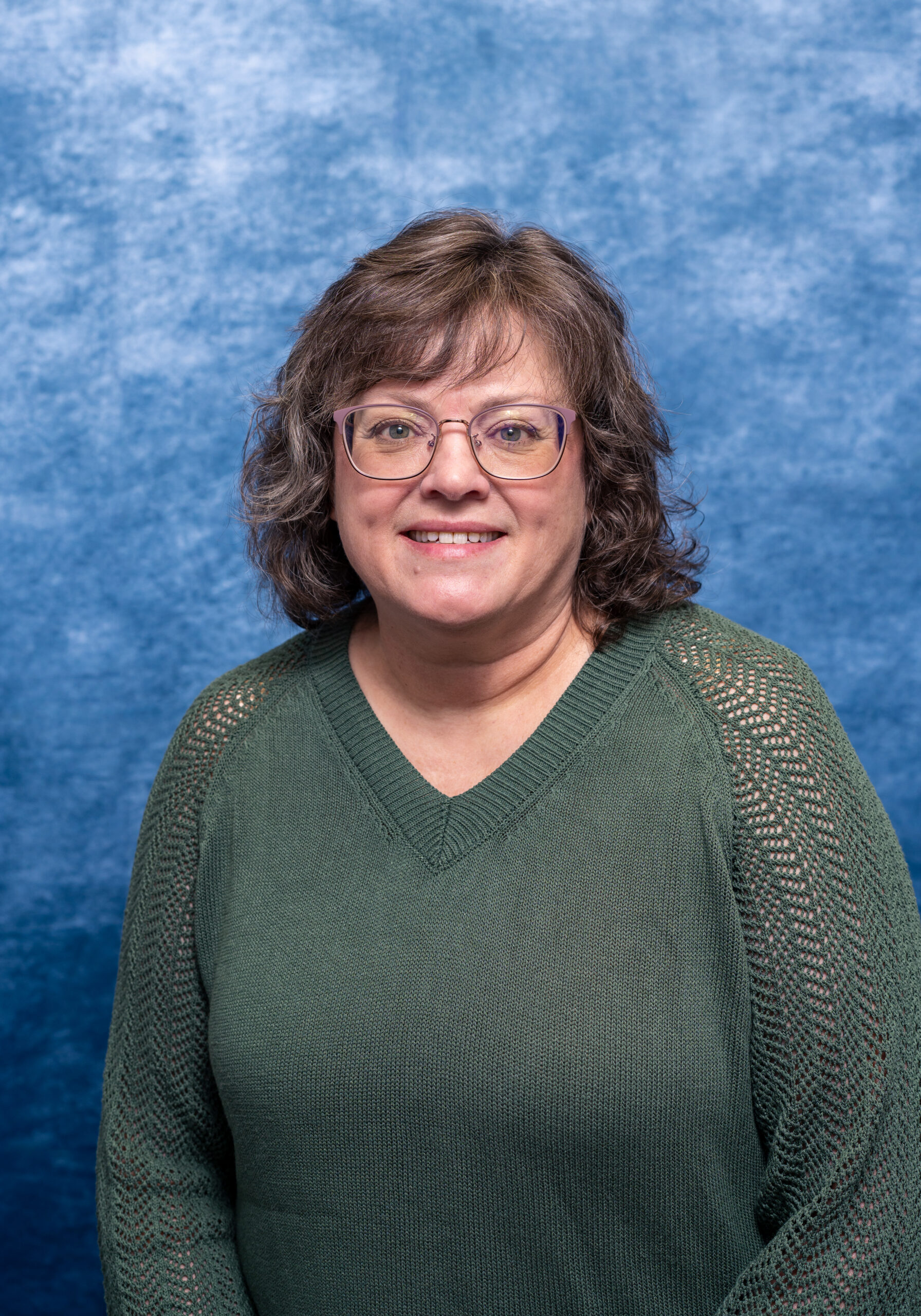 A woman with glasses and curly brown hair smiling in front of a blue textured background.