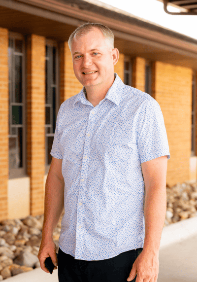 Man smiling outdoors near a brick building, wearing a light blue shirt.