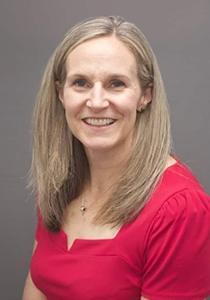 Smiling woman with long blonde hair wearing a red blouse against a gray background.
