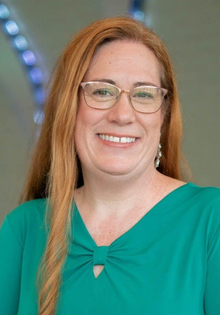 Smile of a woman with red hair and glasses, wearing a teal top, in an indoor setting.