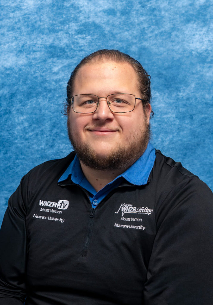 Portrait of Professor Micah Andrew Gerber with glasses and beard, smiling, against blue background.