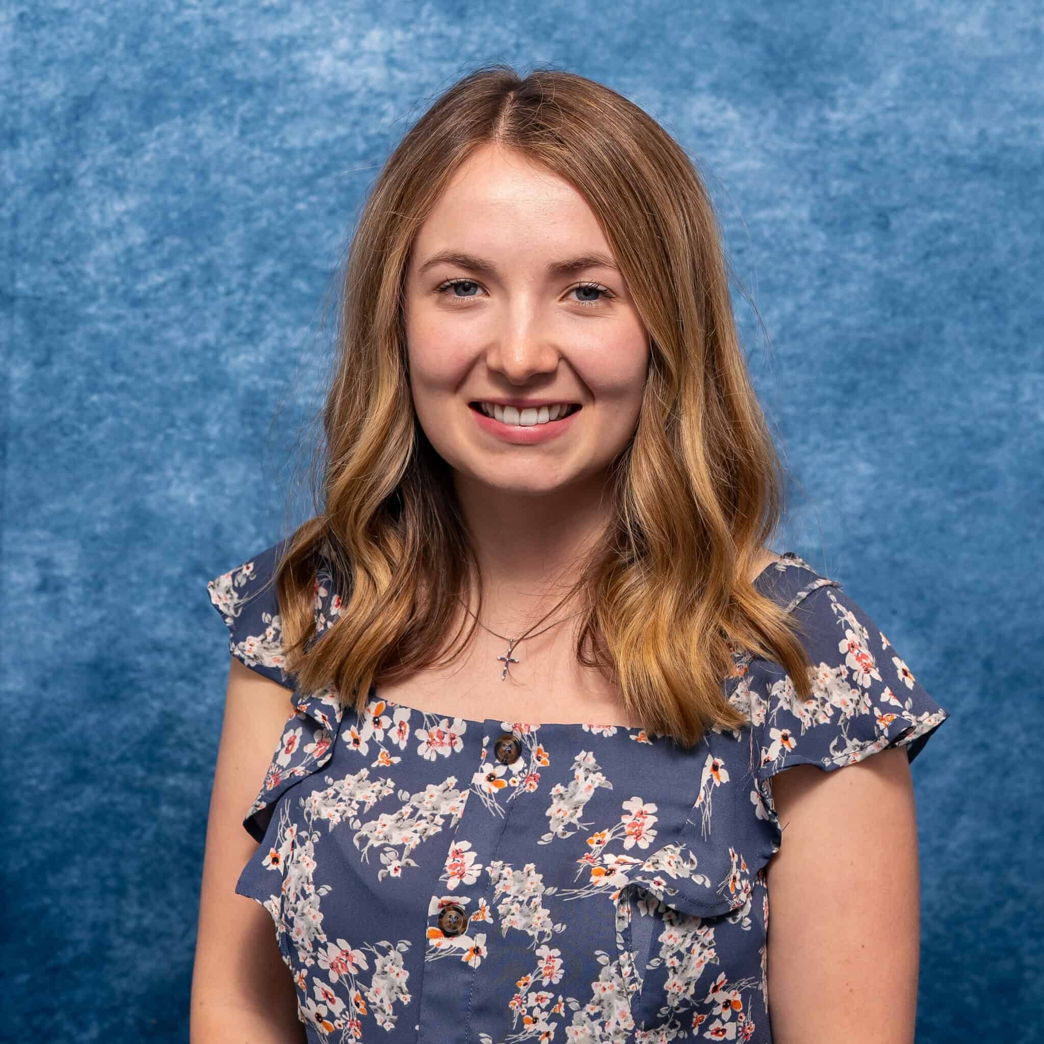 Bright smiling young woman with wavy blonde hair in floral dress against blue textured background.
