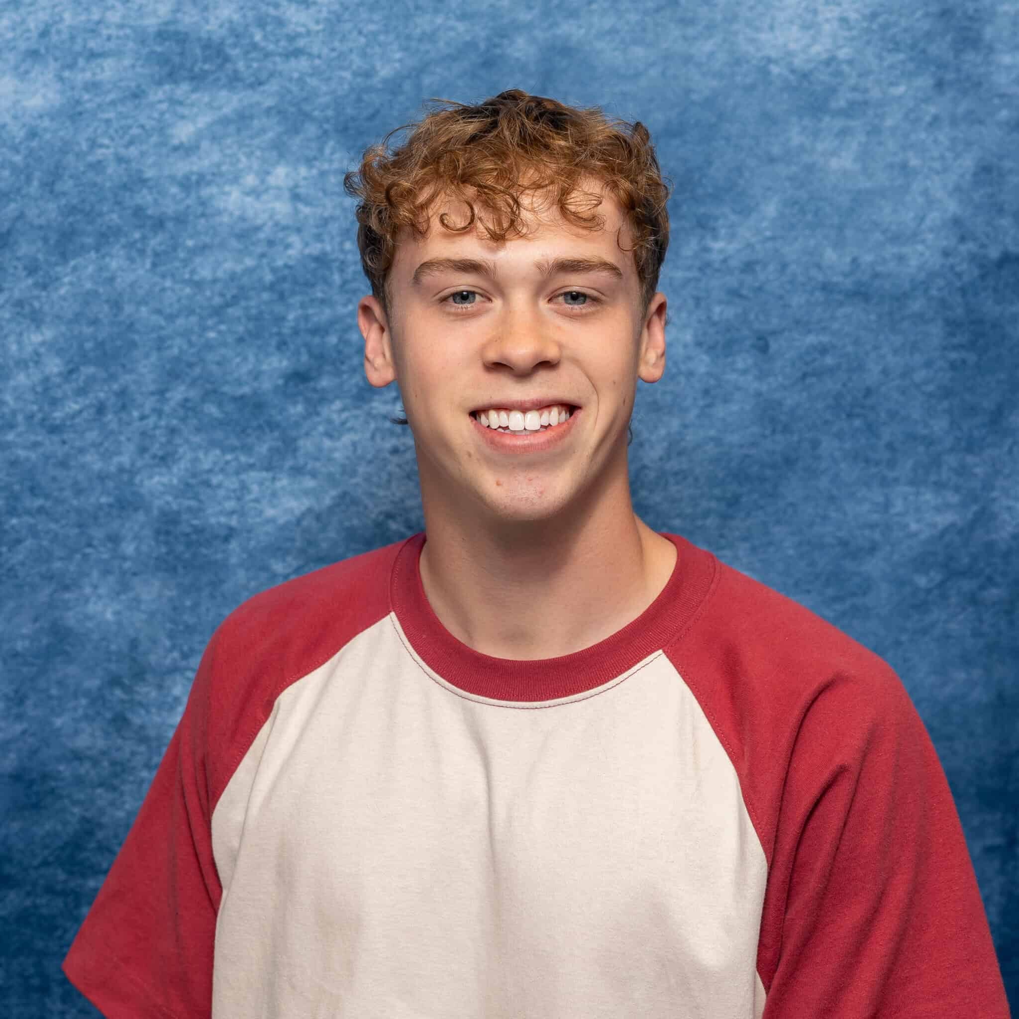 A young man with curly hair smiling in front of a blue textured background.