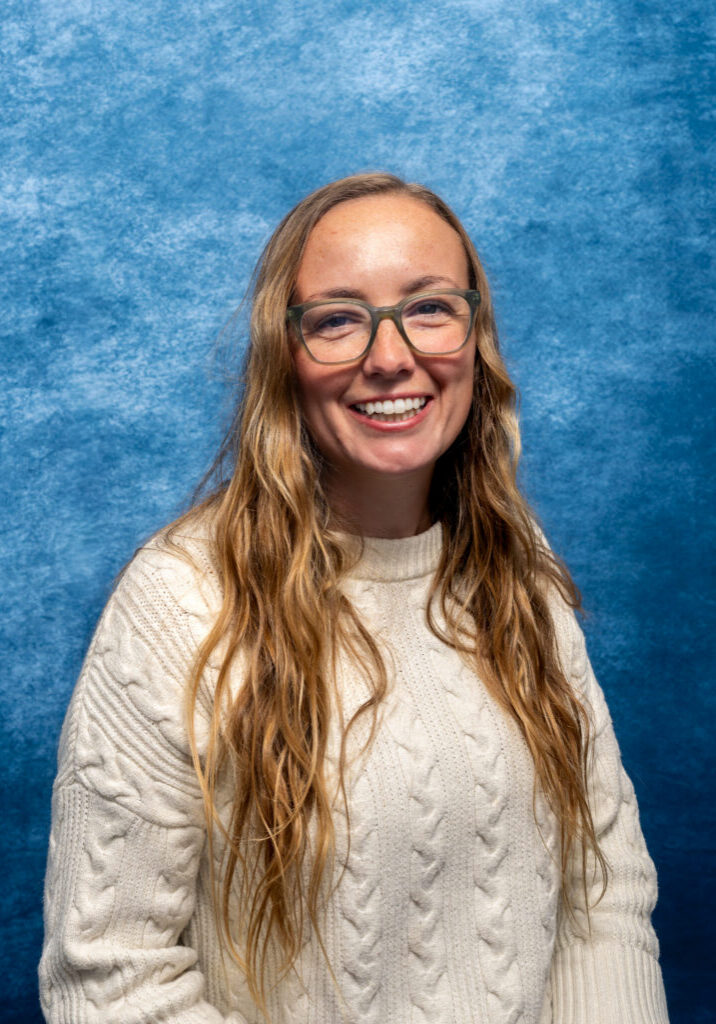 Smiling woman with long wavy hair and glasses against blue background.
