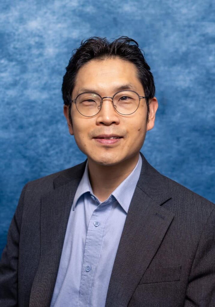 Senior professional man in a suit with glasses, smiling, against a blue textured background.