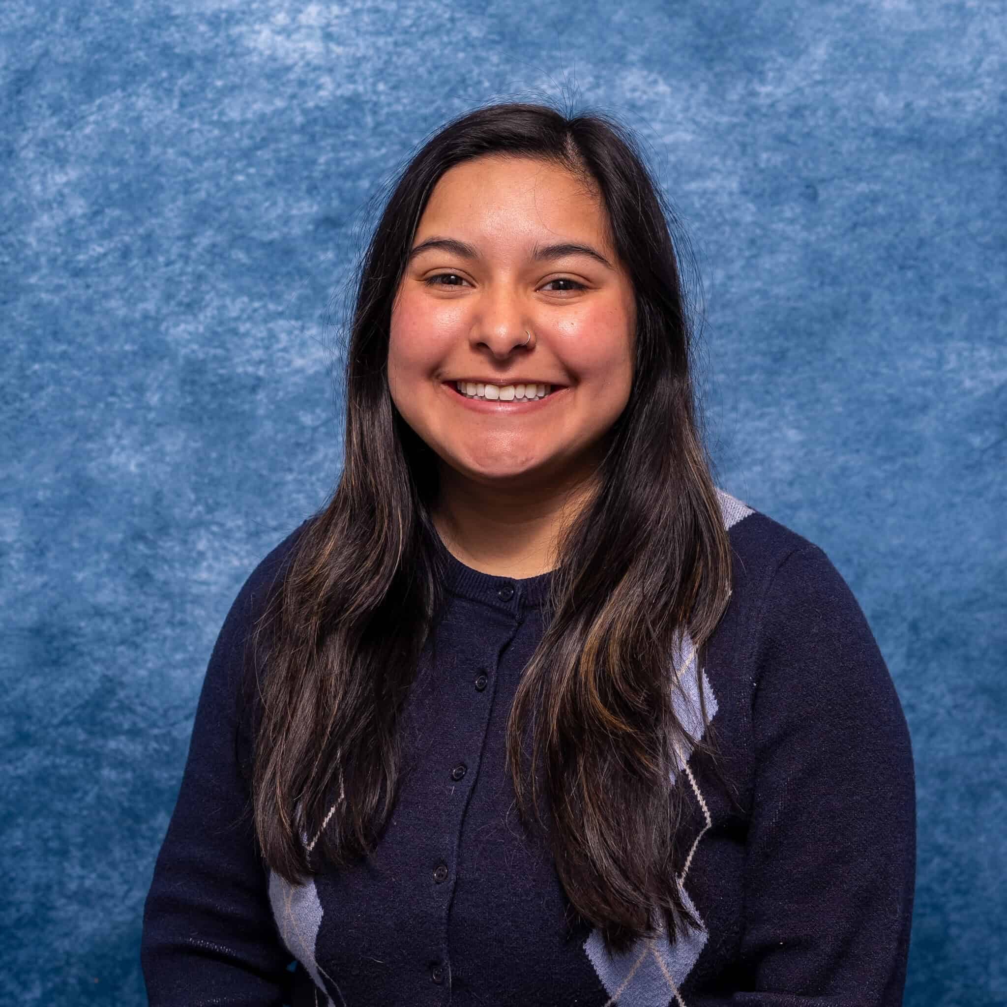 A woman smiling in a navy blue sweater with argyle pattern against a textured blue background.