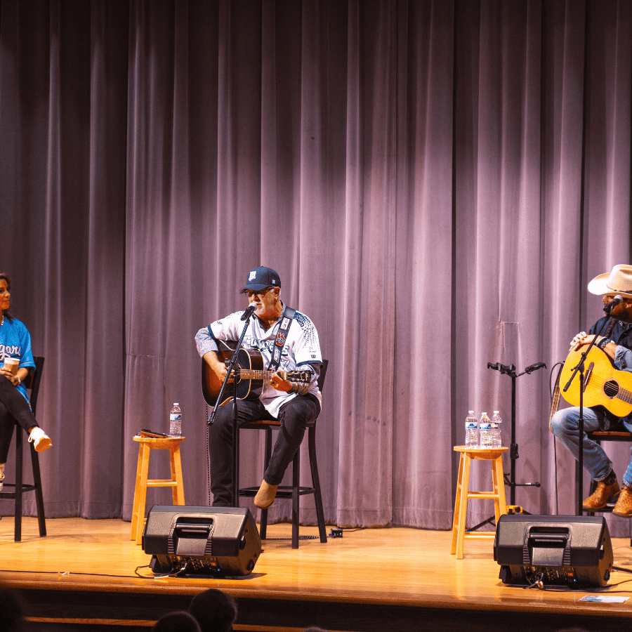 Playing musicians on stage with microphones and water bottles, performing in front of a purple curta.
