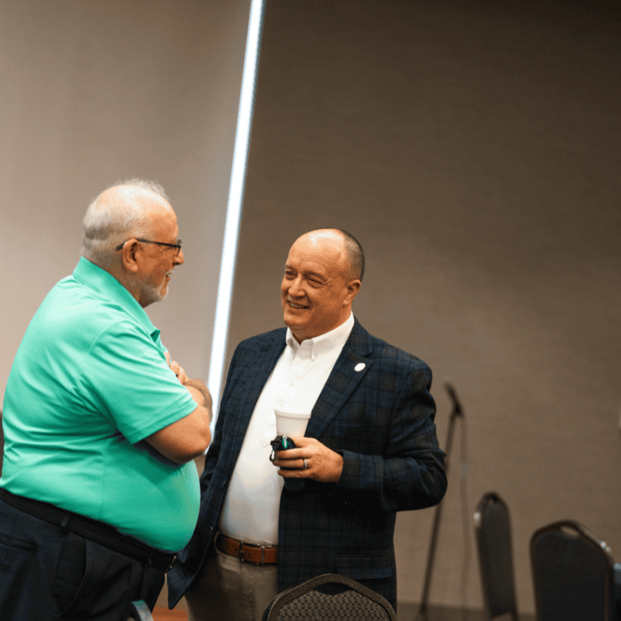 A man in a teal shirt and glasses talking to a man in a dark blazer at a conference.