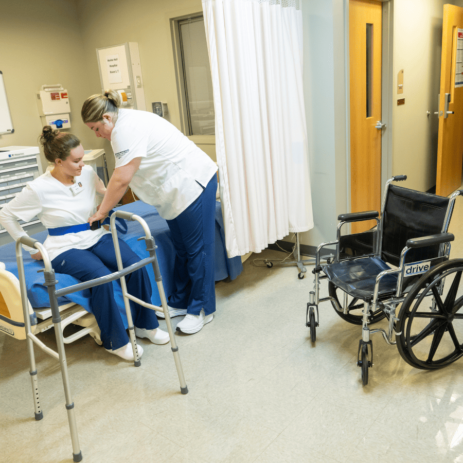Assisting nurse helping patient in hospital room with wheelchair nearby.
