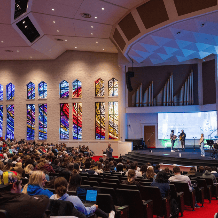 Brightly lit church interior with stained glass windows, congregation seated, and a band performing.