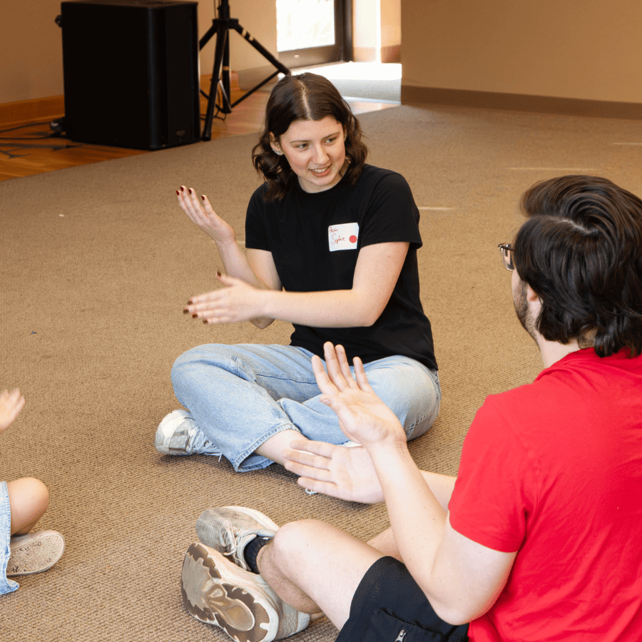 A young woman and a man engage children in a pastoral ministry activity, sitting on the carpeted flo.