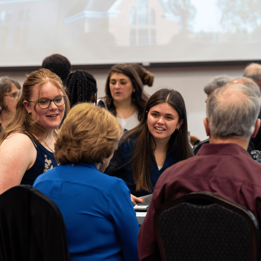 Group of diverse people socializing at a formal event with tables and a large screen.