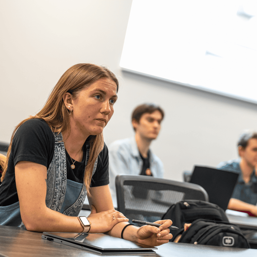 Woman attentively listening during a classroom lecture with students in the background.