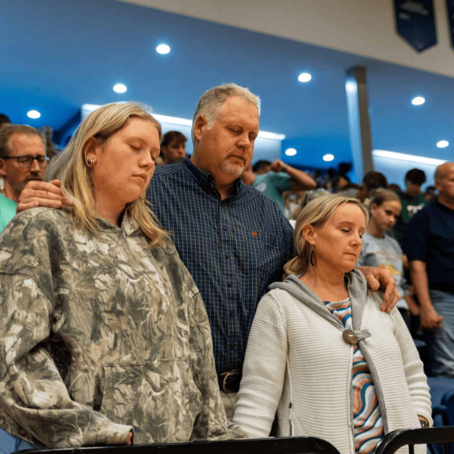 Parents praying with children during church service, eyes closed, in a congregation setting.
