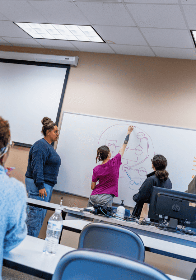 A woman writes on a whiteboard during a classroom presentation with students seated at desks.