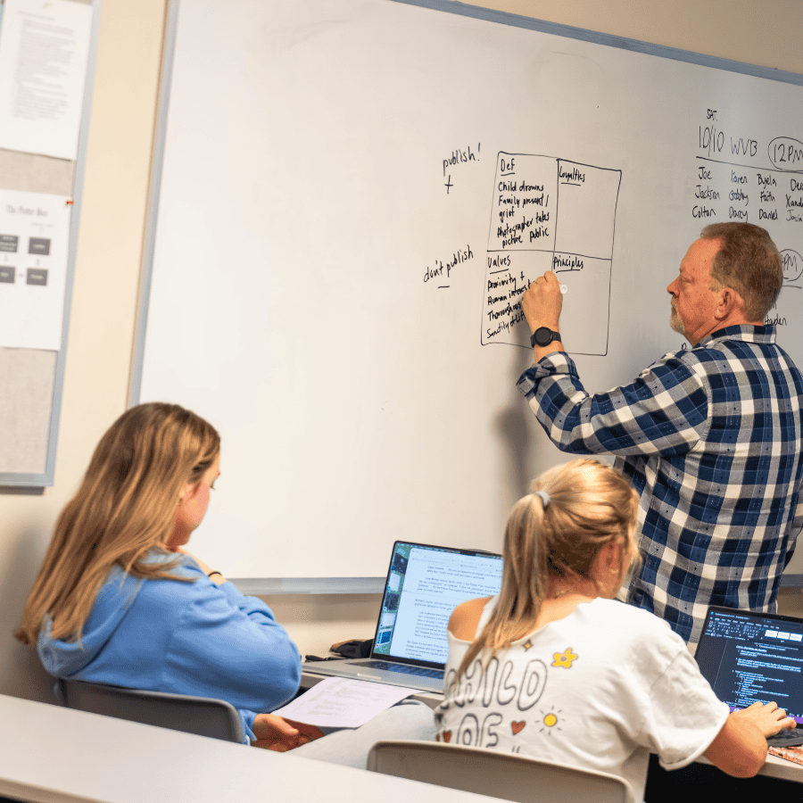 Teacher explaining concepts on whiteboard to students in a classroom.