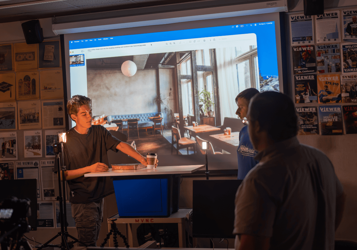 Young man giving a presentation to two people in a classroom with a large screen.